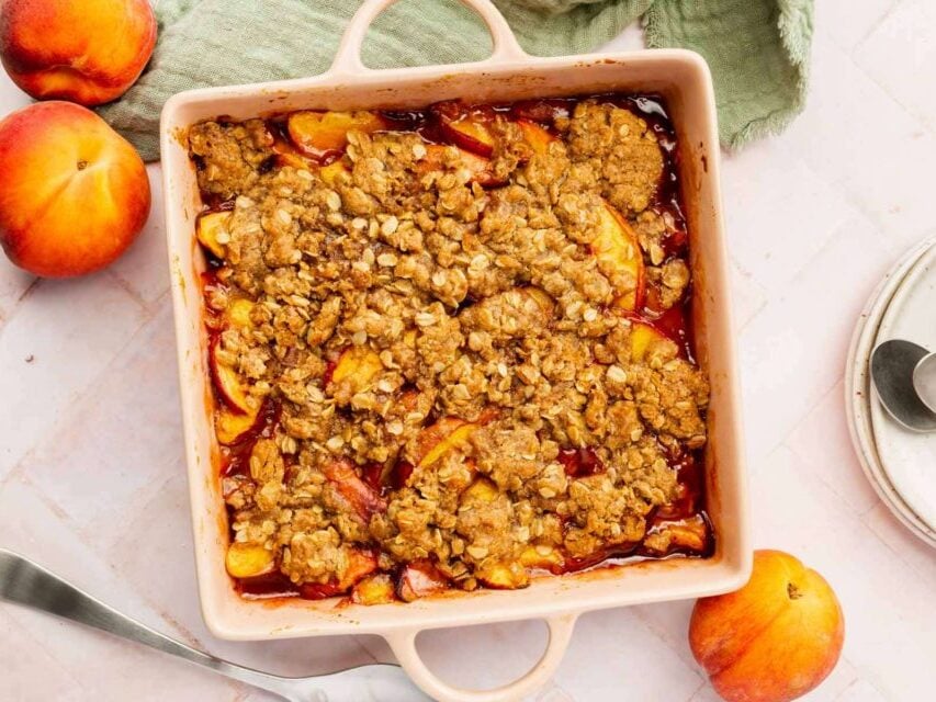 An overhead view of gluten-free vegan peach crisp in a square baking dish with fresh peaches on the table.
