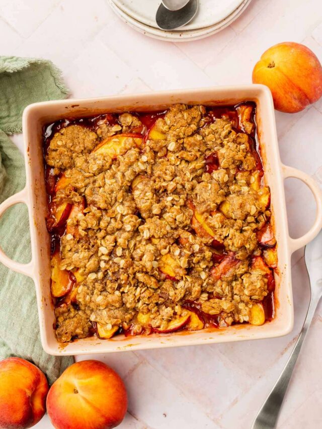 An overhead view of gluten-free vegan peach crisp in a square baking dish with fresh peaches on the table.
