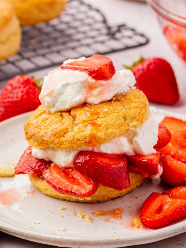 A gluten-free strawberry shortcake on a plate topped with fresh whipped cream with whole strawberries in the background.