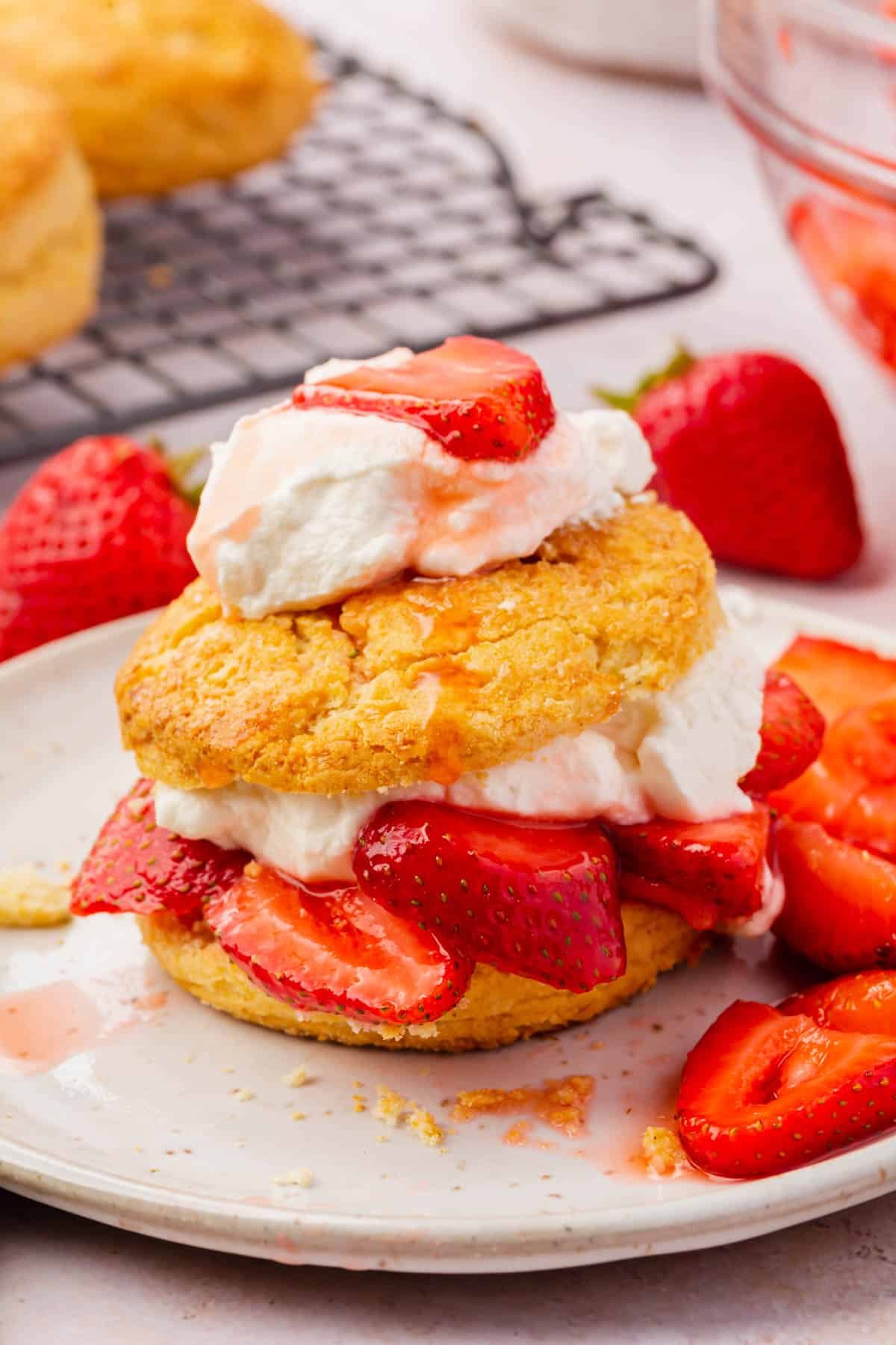 A gluten-free strawberry shortcake on a plate topped with fresh whipped cream with whole strawberries in the background.