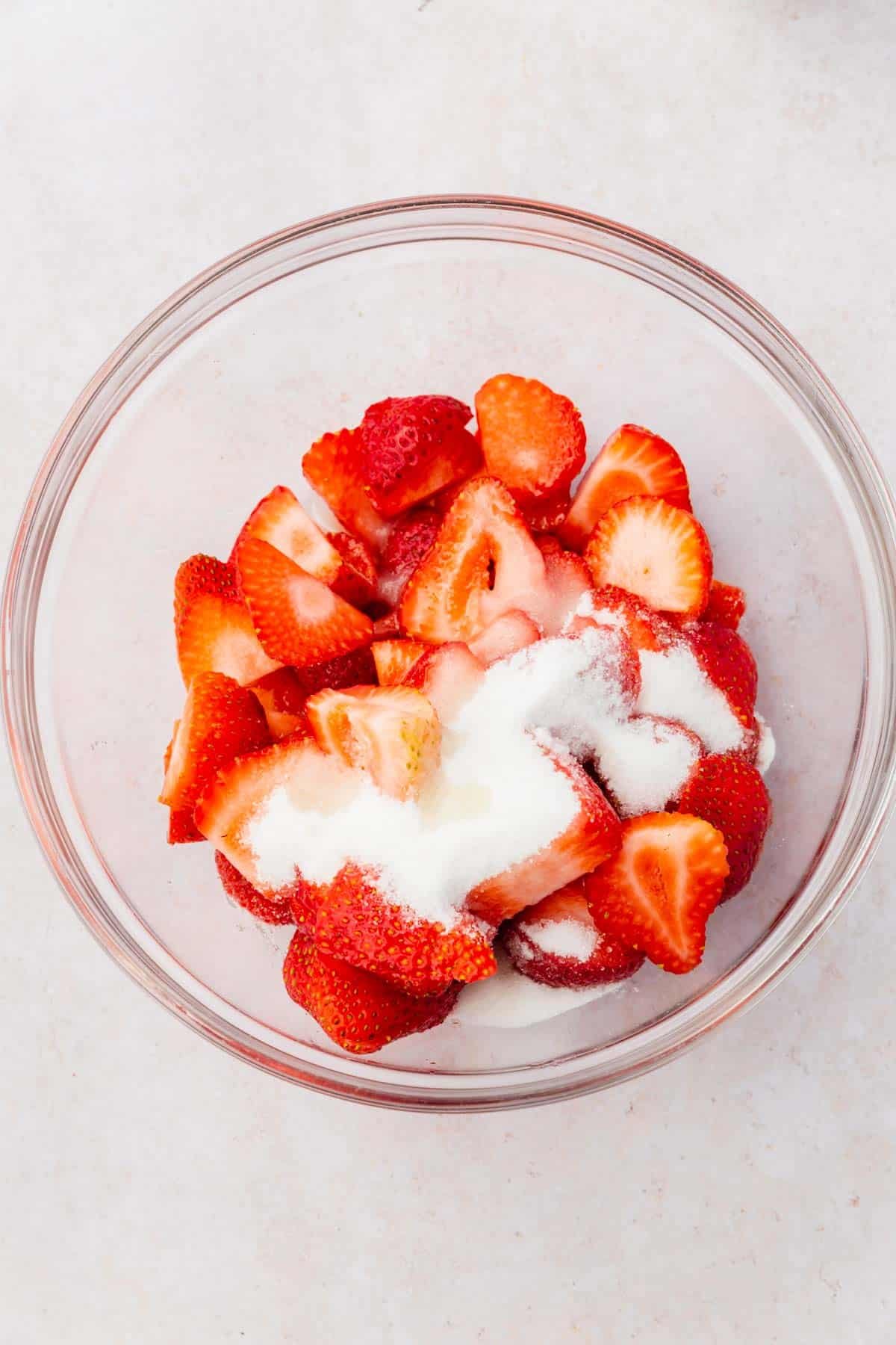 An overhead view of a mixing bowl with sliced strawberries, lemon juice and granulated sugar before mixing.