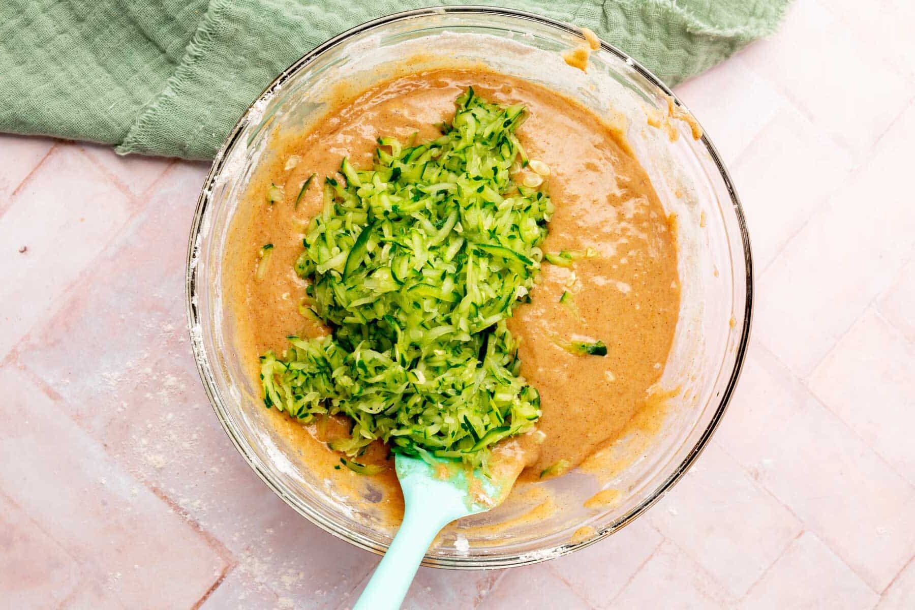 A glass bowl with grated zucchini on top of mixed wet ingredients to make gluten-free zucchini bread.