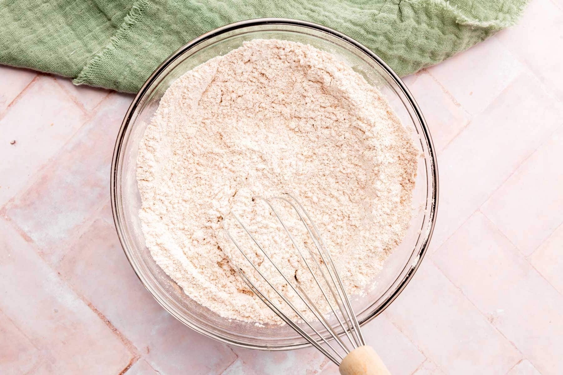 A large glass mixing bowl with dry ingredients for zucchini bread with a whisk.