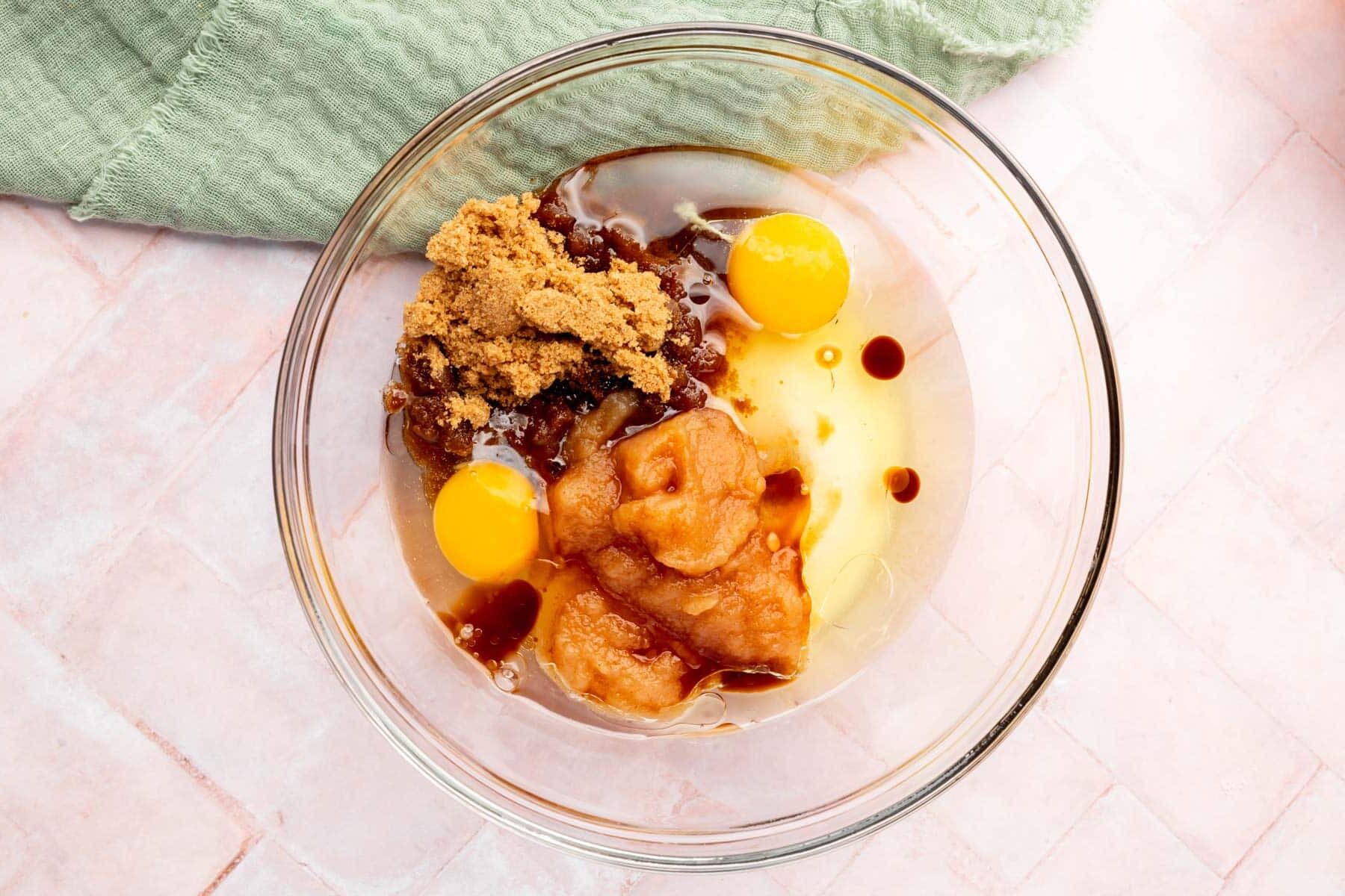 A large glass bowl filled with oil, eggs, vanilla extract, brown sugar, and applesauce.