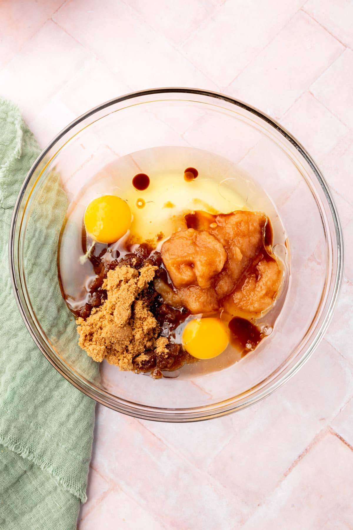 A large glass bowl filled with oil, eggs, vanilla extract, brown sugar, and applesauce.