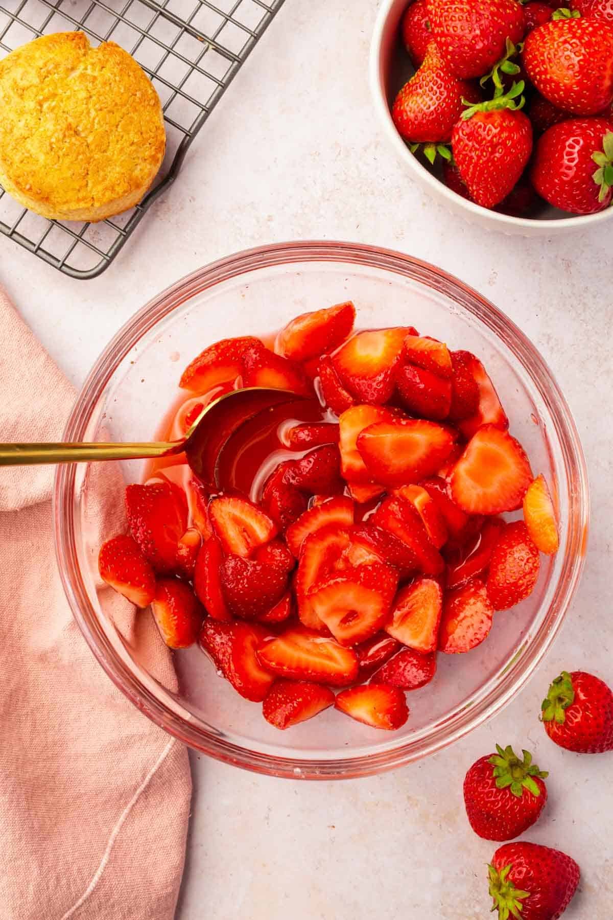 An overhead view of a glass bowl of macerated strawberries with a spoon, a bowl of whole strawberries and a cooling rack with gluten-free biscuits peaking in.