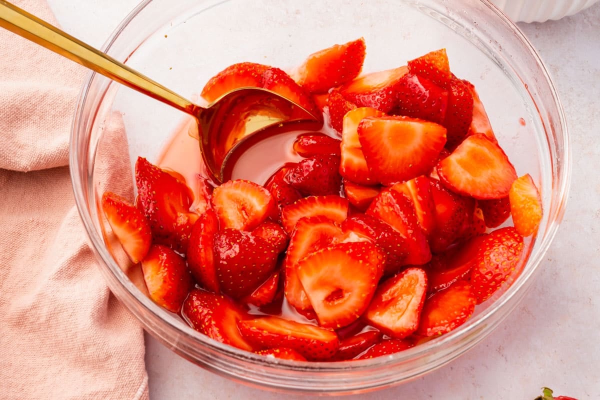A glass mixing bowl with macerated strawberries and a gold spoon.