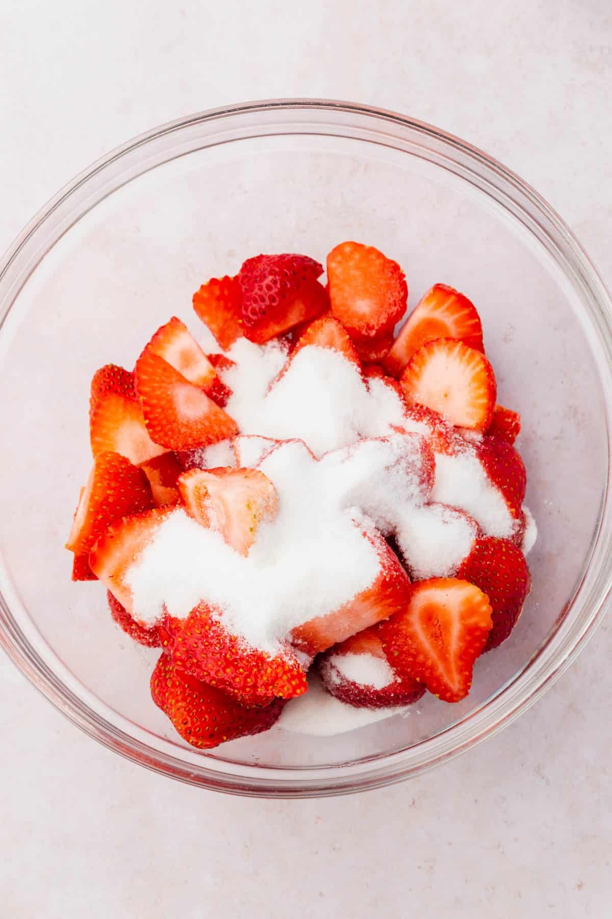 A glass mixing bowl with sliced strawberries and granulated sugar on top that have not been mixed.