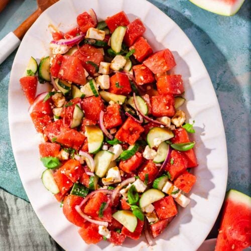 A white scalloped oval platter with watermelon feta salad topped with cucumber and basil with large slices of watermelon on the side.