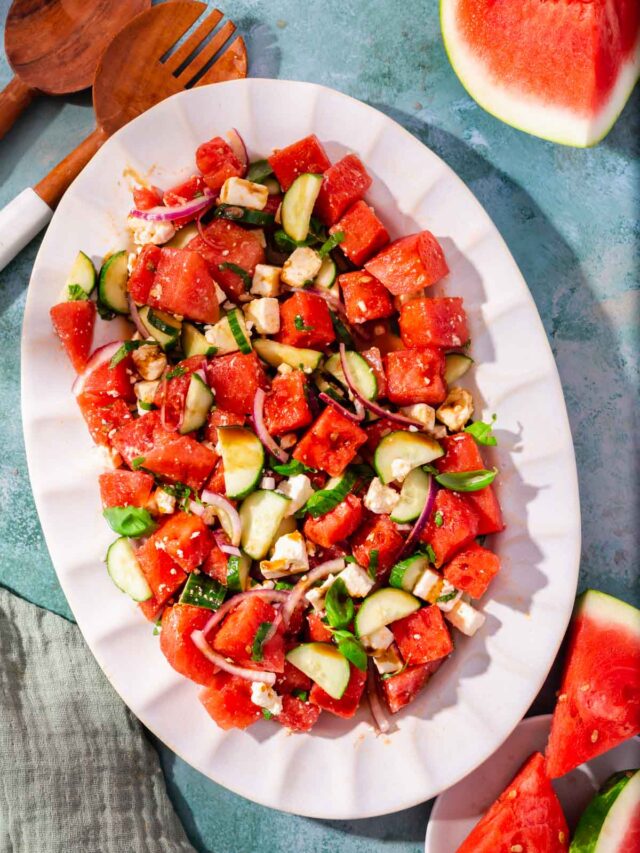 A white scalloped oval platter with watermelon feta salad topped with cucumber and basil with large slices of watermelon on the side.