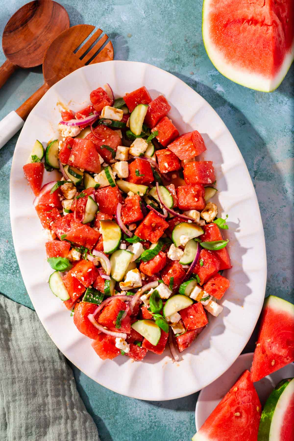 A white scalloped oval platter with watermelon feta salad topped with cucumber and basil with large slices of watermelon on the side.