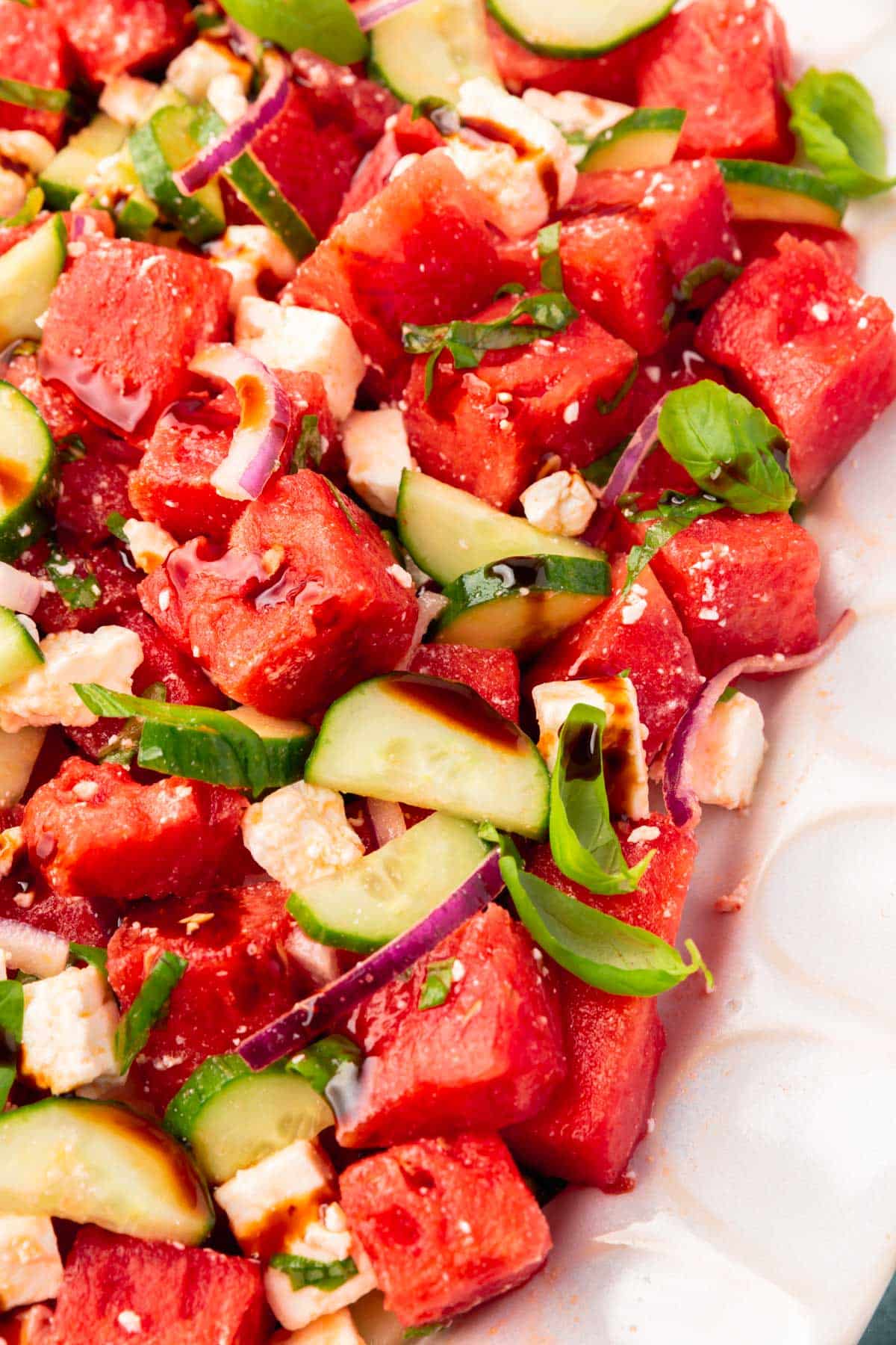 A closeup of watermelon salad with cucumber, red onion, feta and basil on a white scalloped oval platter.