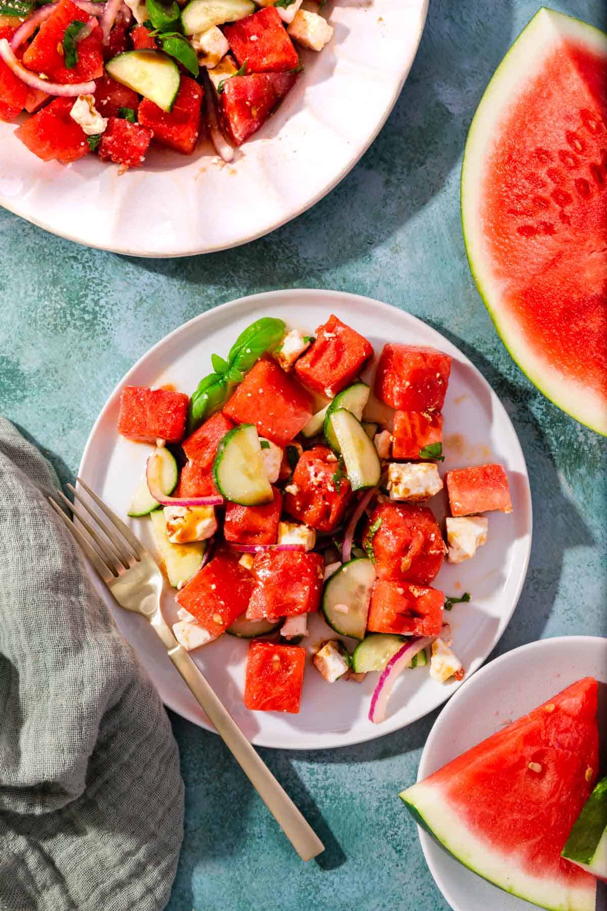 A salad plate with watermelon feta cucumber salad on it with large pieces of watermelon on teh side.