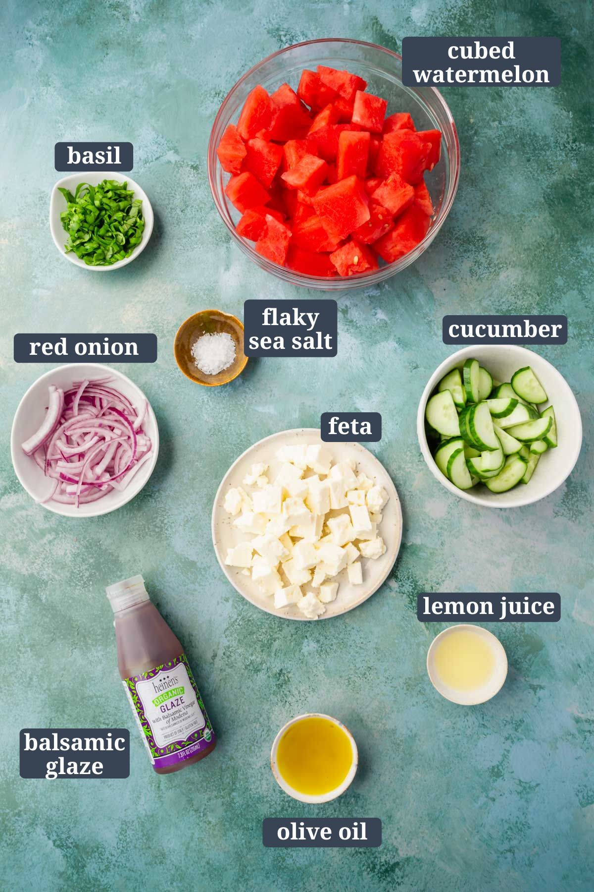 Small bowls of ingredients on a green table to make watermelon cucumber feta salad with basil with text overlays over each ingredient.