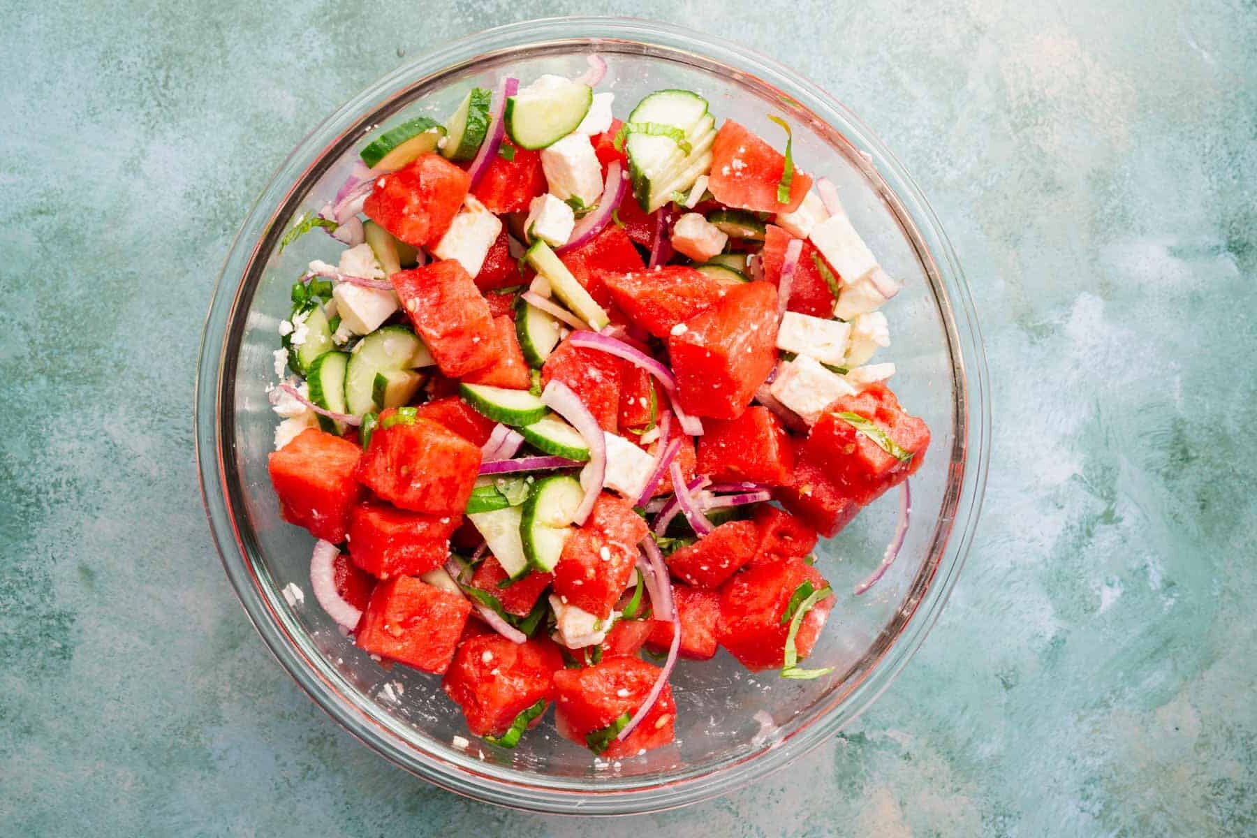 A mixing bowl with a watermelon salad tossed with feta, sliced cucumber, sliced red onion, sliced basil, lemon juice and olive oil in it.