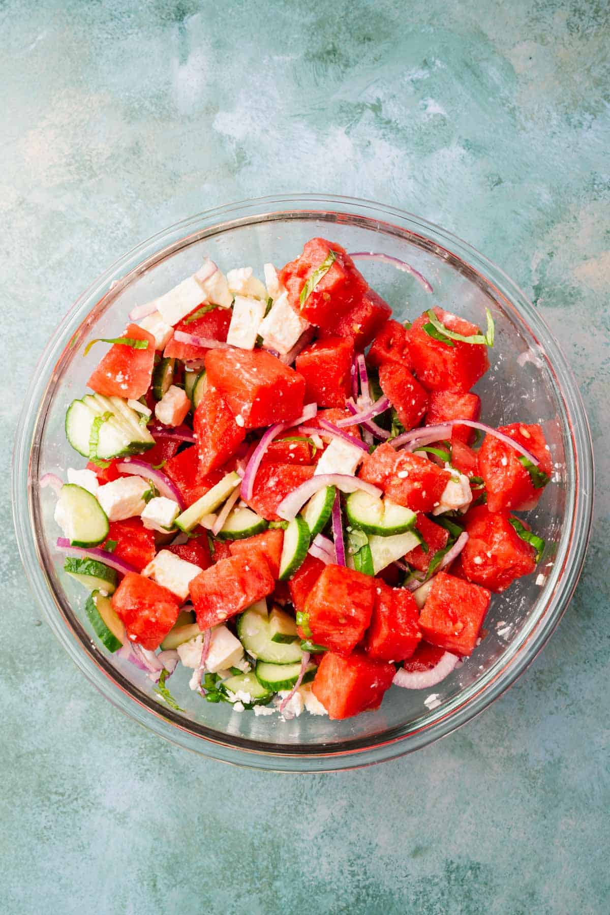 A mixing bowl with a watermelon salad tossed with feta, sliced cucumber, sliced red onion, sliced basil, lemon juice and olive oil in it.