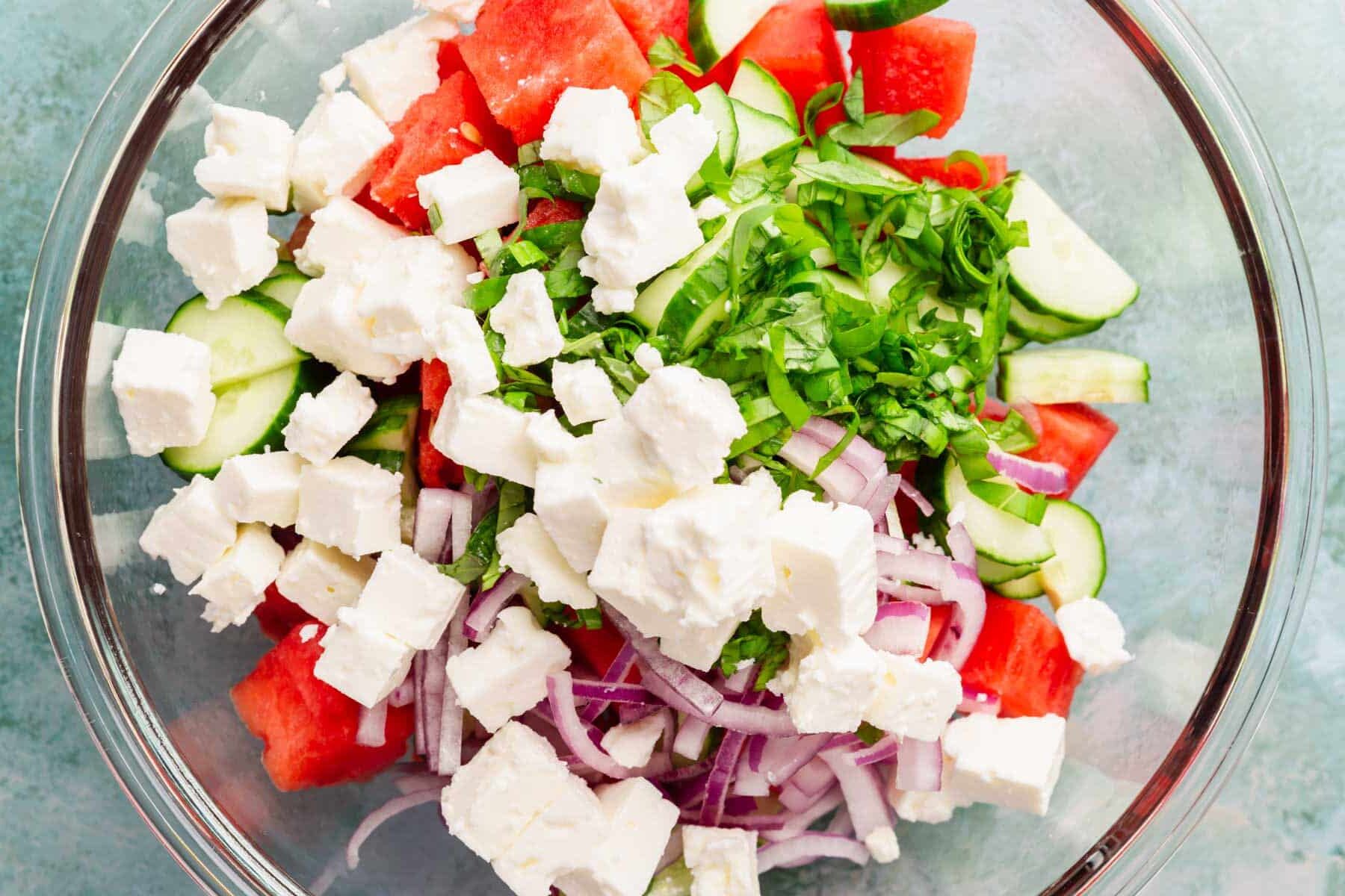 A mixing bowl with diced watermelon, basil, sliced red onion, sliced cucumber and cubed feta cheese in it.