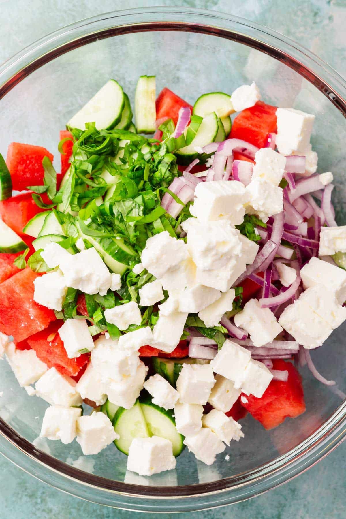 A mixing bowl with diced watermelon, basil, sliced red onion, sliced cucumber and cubed feta cheese in it.