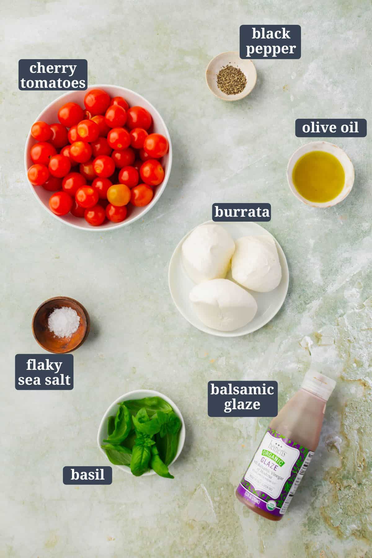 An overhead view of small bowls of ingredents on a table to make burrata caprese salad with text overlays over each item.