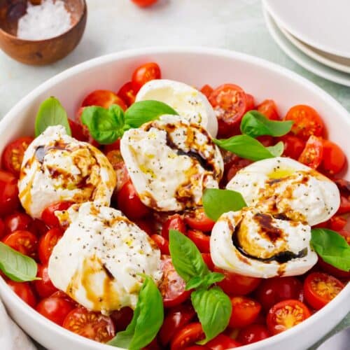 A shallow bowl of burrata caprese salad with cherry tomatoes in the background.