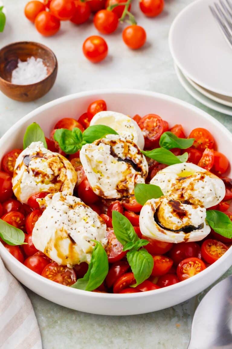 A shallow bowl of burrata caprese salad with cherry tomatoes in the background.