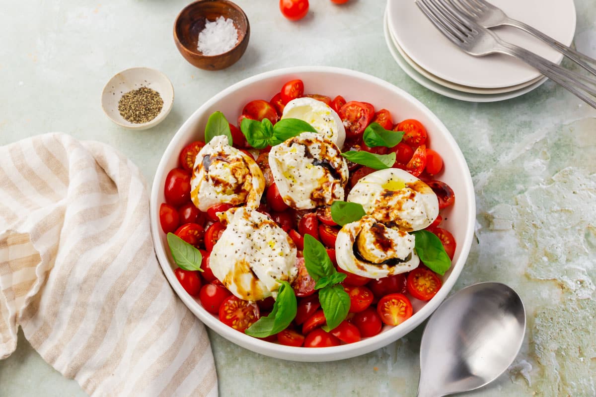 A shallow bowl of burrata caprese salad surrounded by small bowls of salt and pepper.