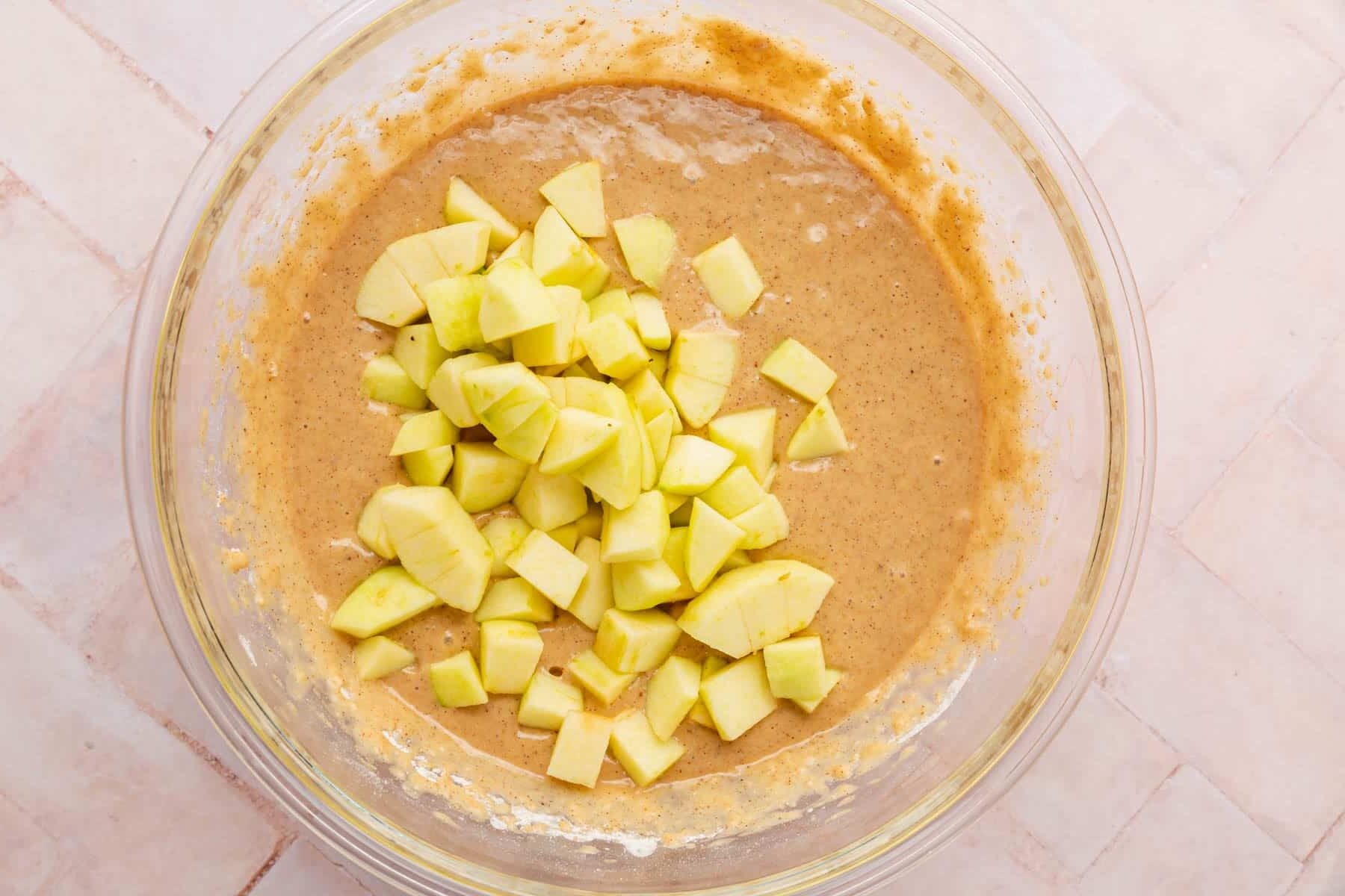 A glass mixing bowl with gluten-free spiced cake batter topped with diced apples in it.