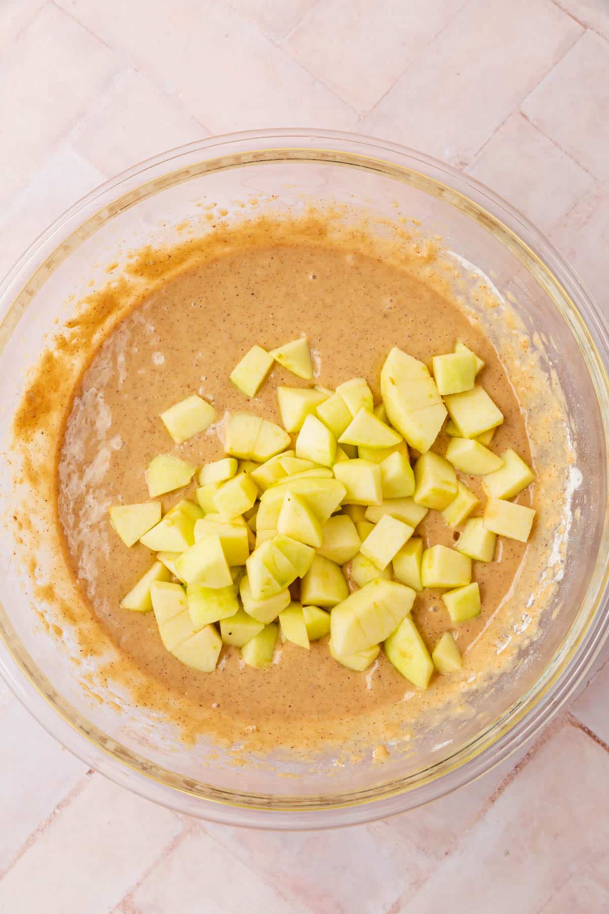 A glass mixing bowl with gluten-free spiced cake batter topped with diced apples in it.