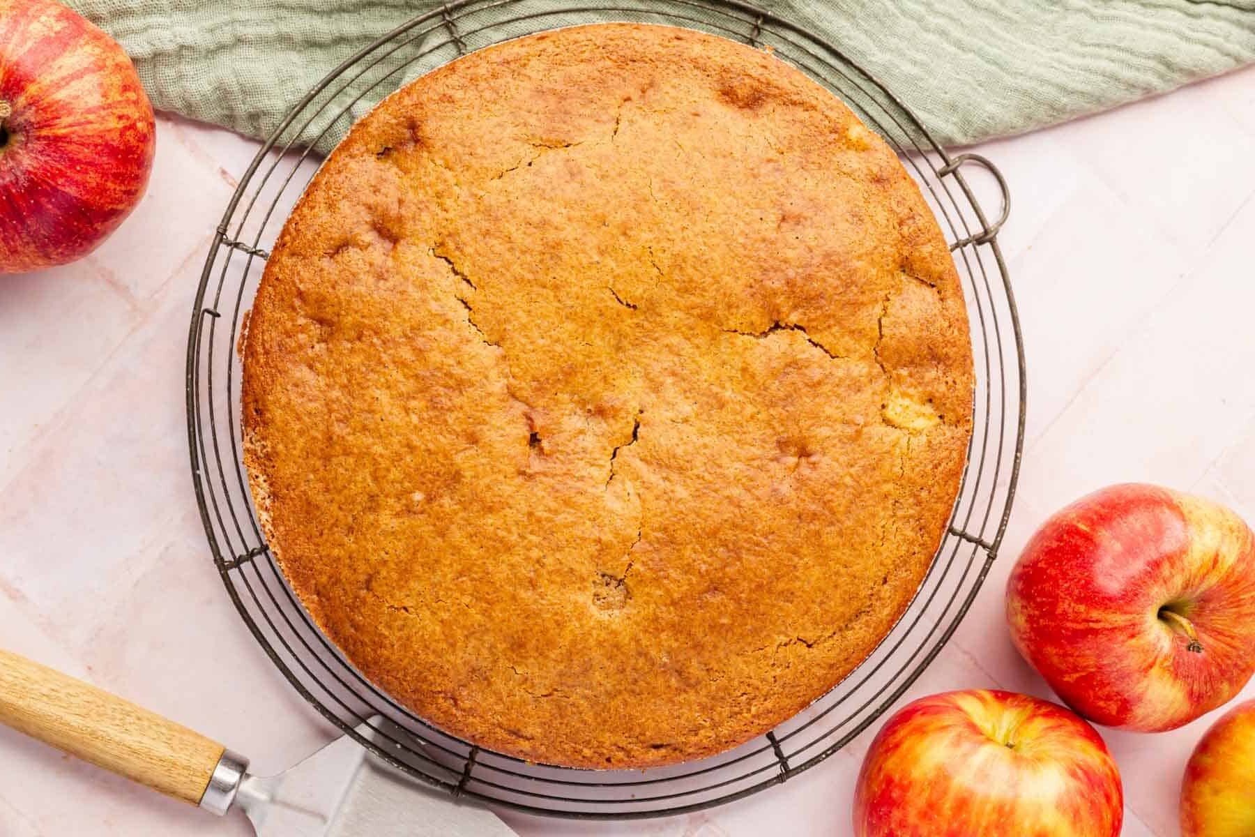 An overhead view of a round gluten-free apple cake on a cooling rack surrounded by whole apples and a green linen napkin.