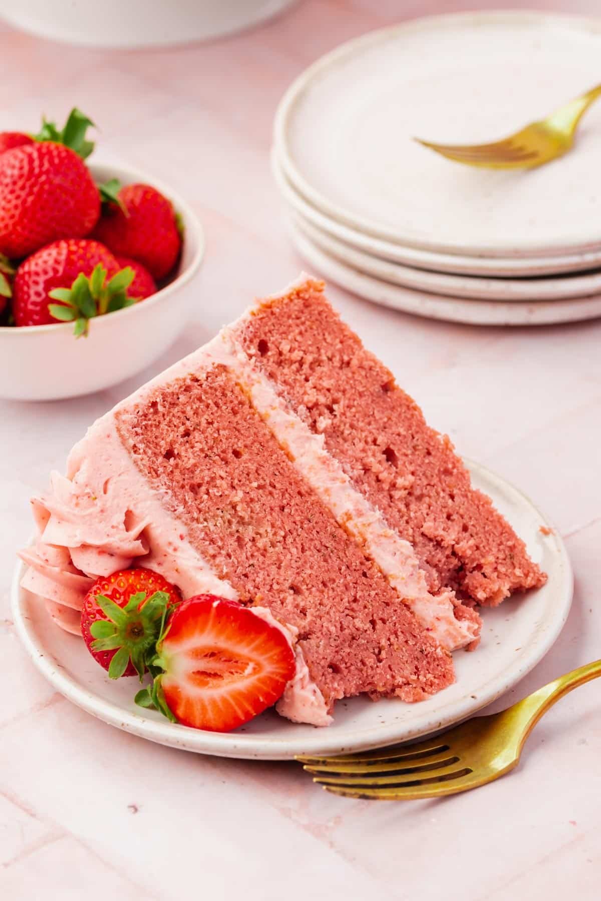A slice of gluten-free strawberry cake on a plate with fresh strawberries next to a gold fork.