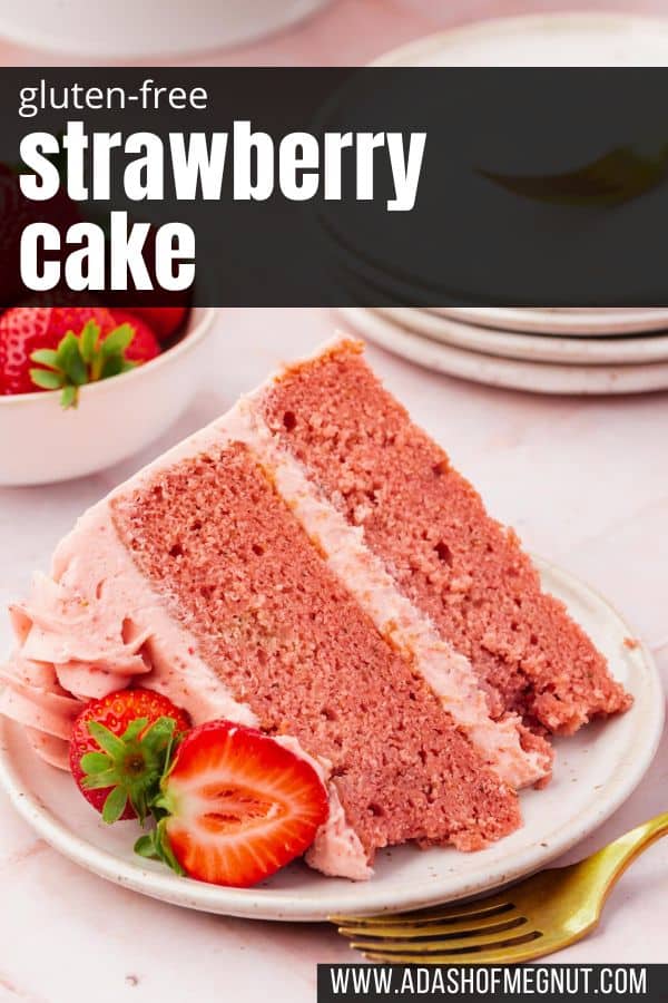 A slice of strawberry cake with strawberry frosting on a dessert plate with fresh strawberry halves and a gold fork.