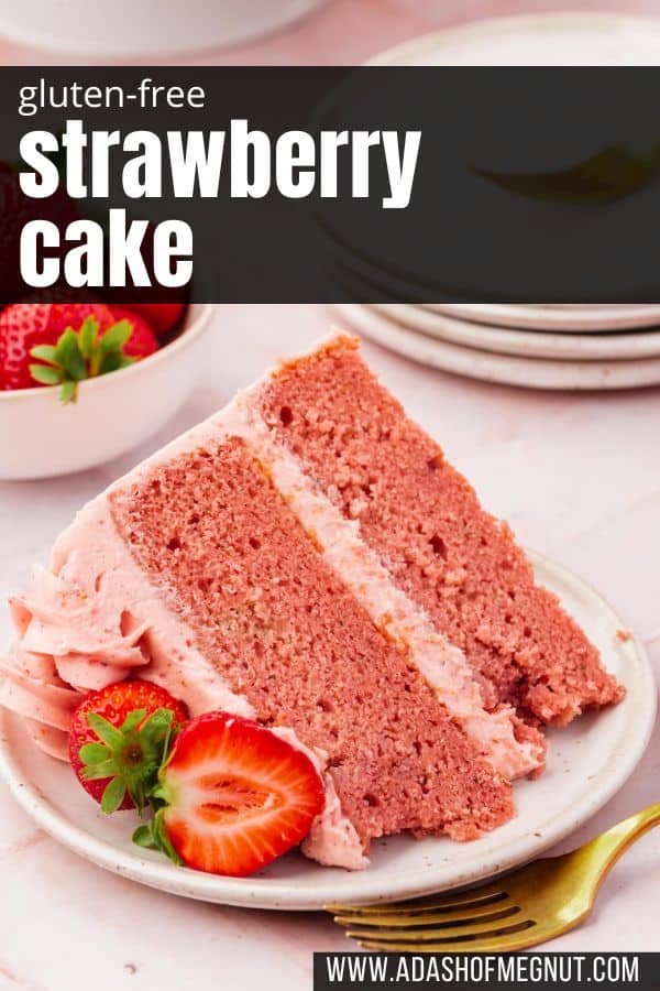 A slice of strawberry cake with strawberry frosting on a dessert plate with fresh strawberry halves and a gold fork.