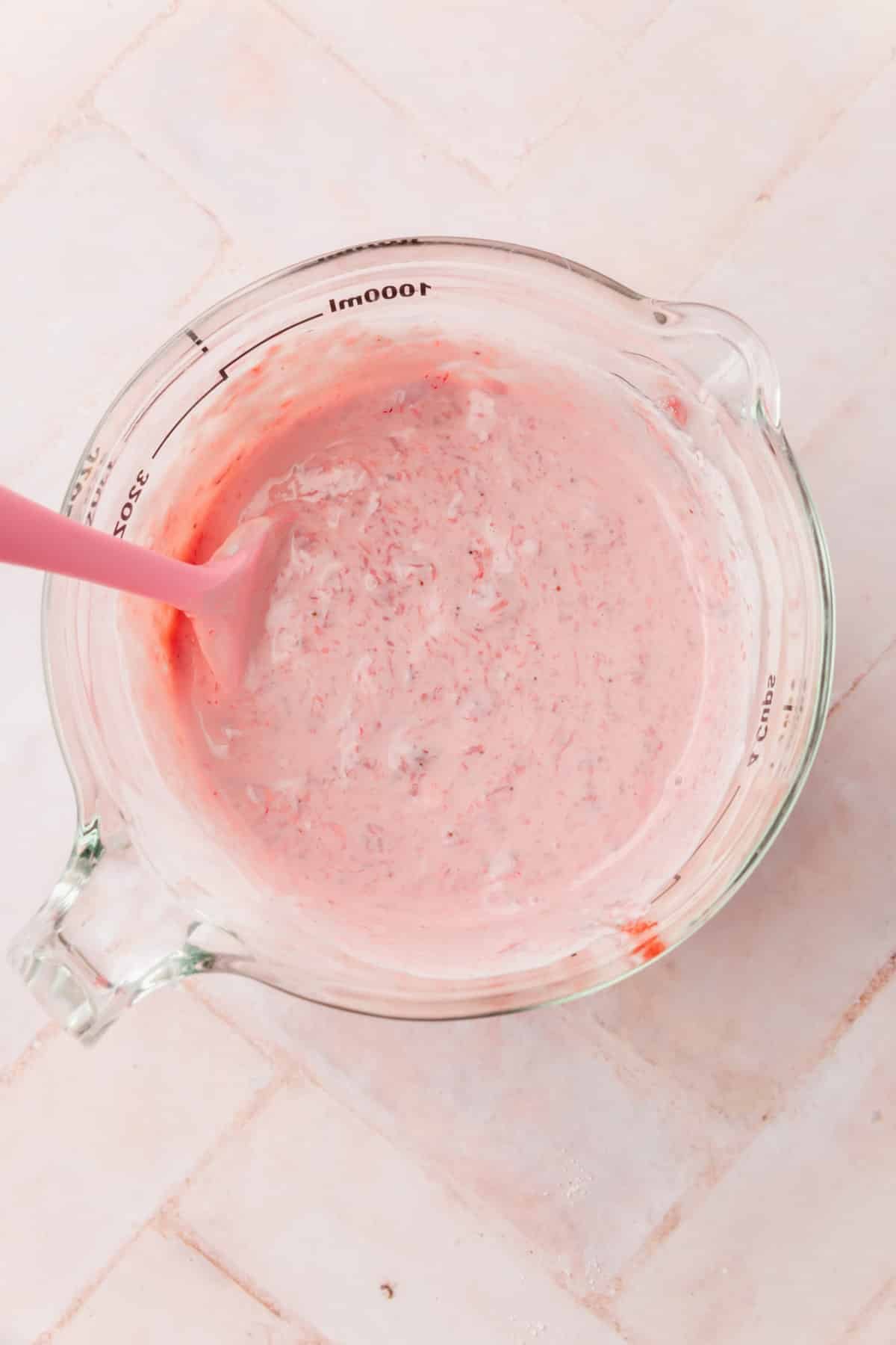 A glass measuring cup with a strawberry sour cream mixture in it with a pink spatula.