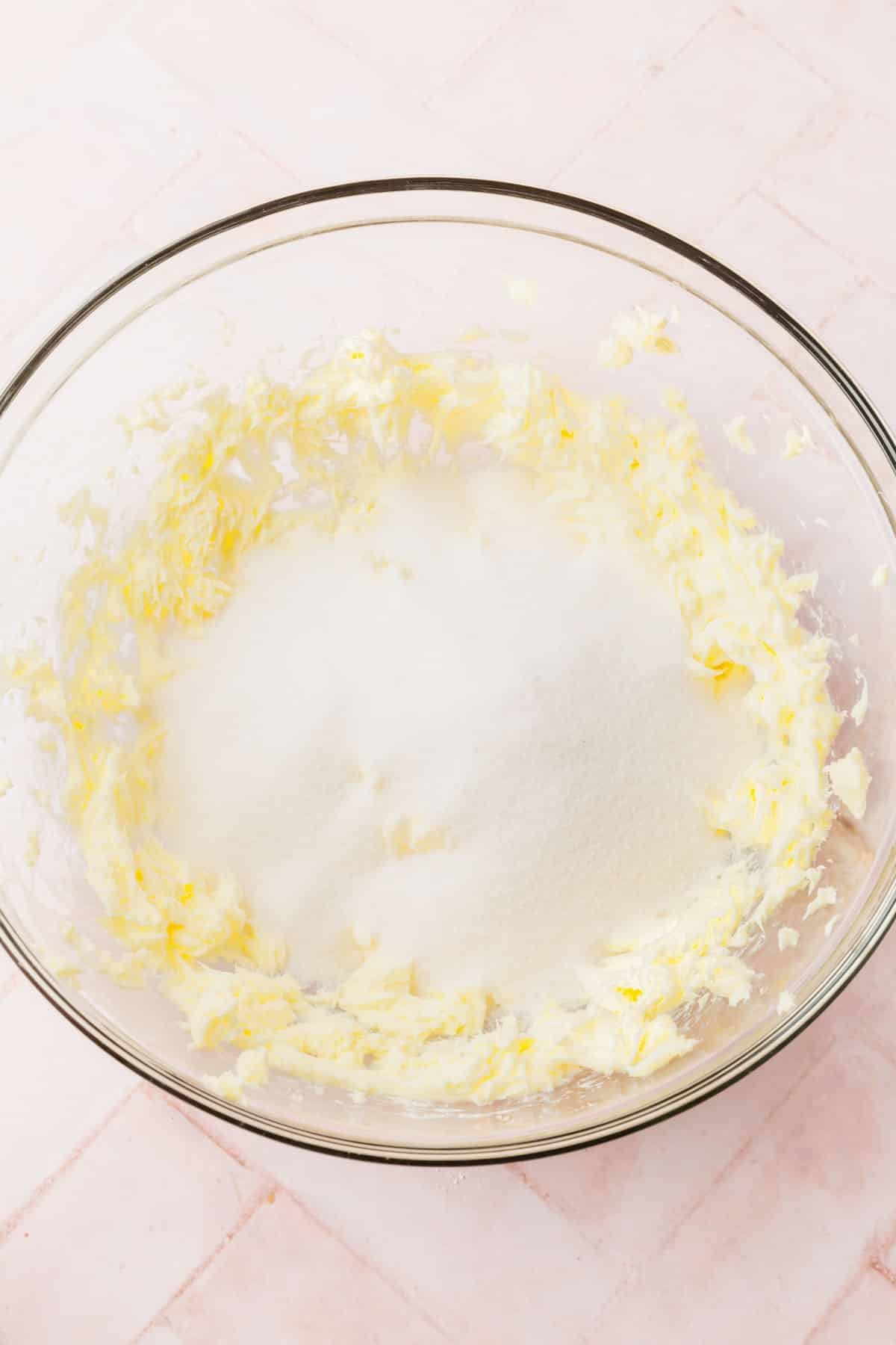 A glass mixing bowl with creamed butter topped with granulated sugar before mixing.