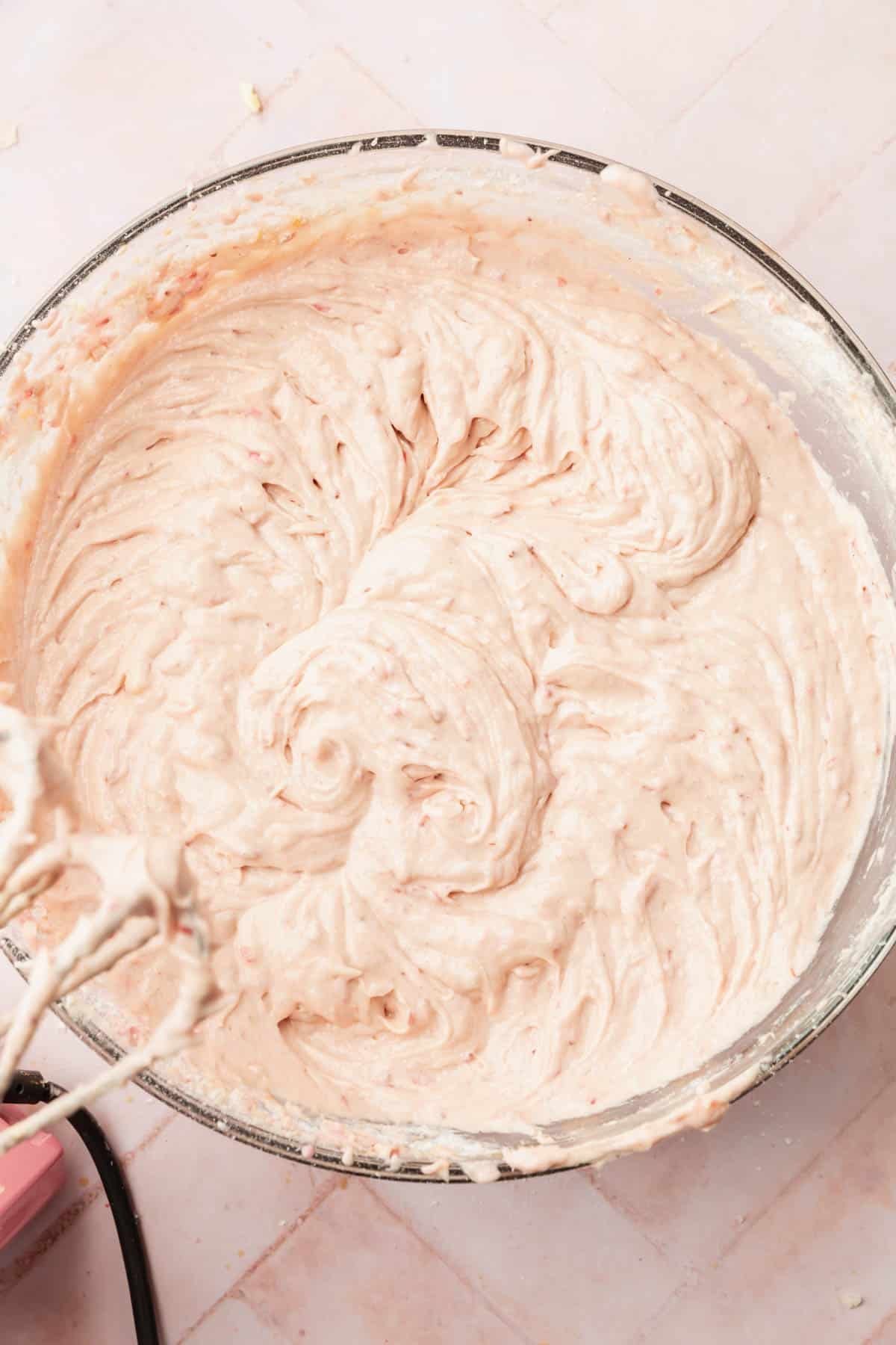 An overhead view of a large mixing bowl with strawberry cake batter in it with electric beaters off to the side.