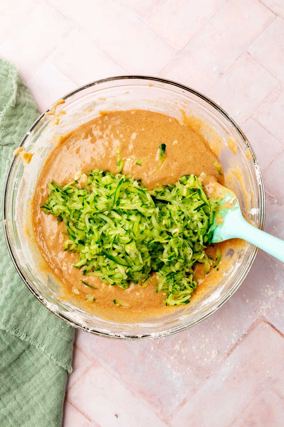 A glass mixing bowl with shredded zucchini being mixed into gluten-free muffin batter.