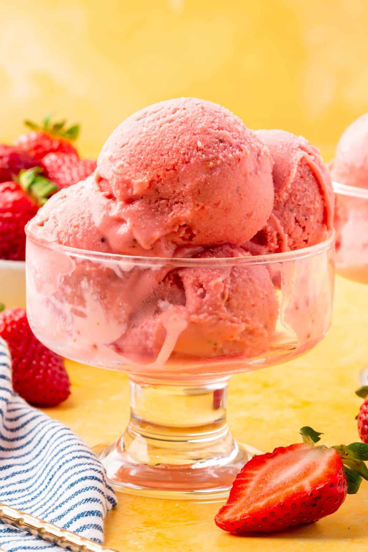 A glass footed bowl with scoops of strawberry froyo in it on a yellow table.