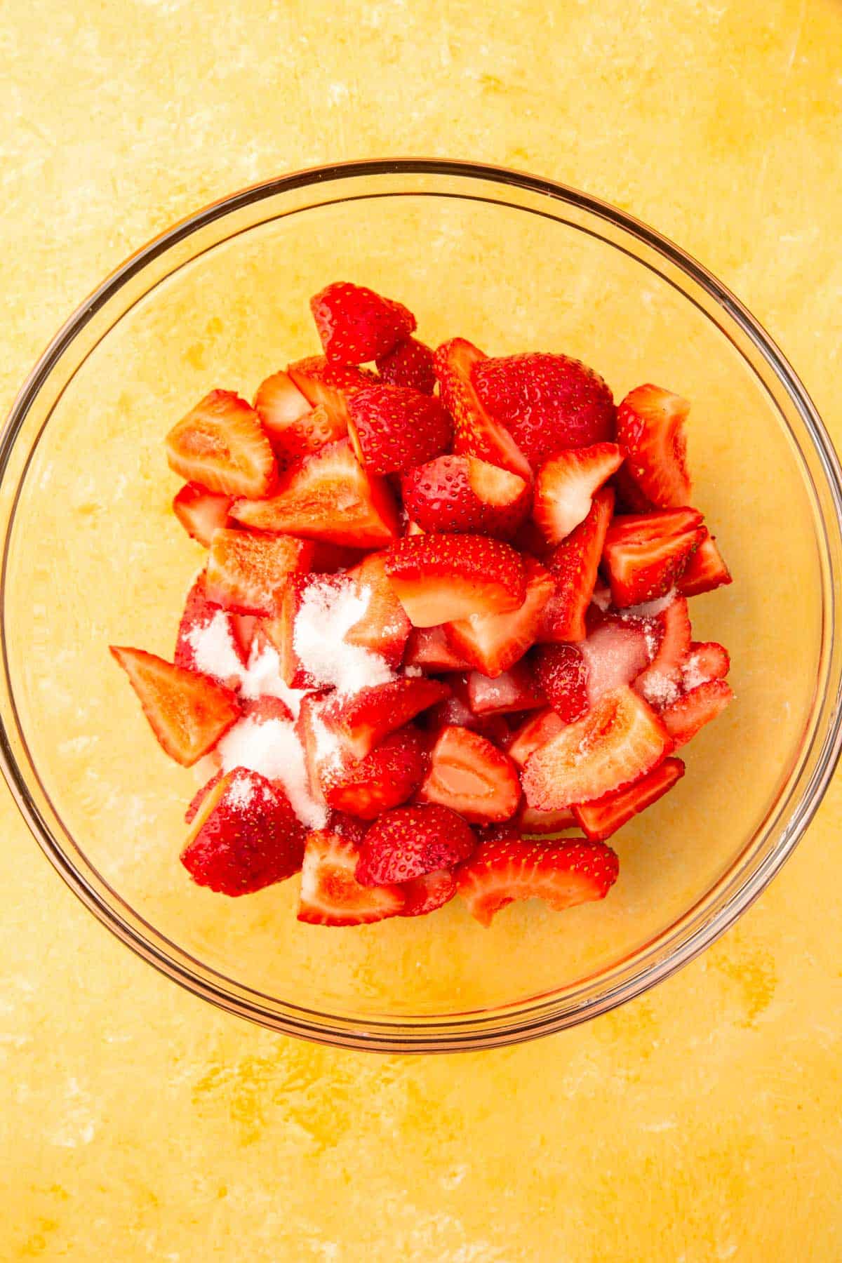 A glass bowl with sliced strawberries, granulated sugar, and lemon juice in it.