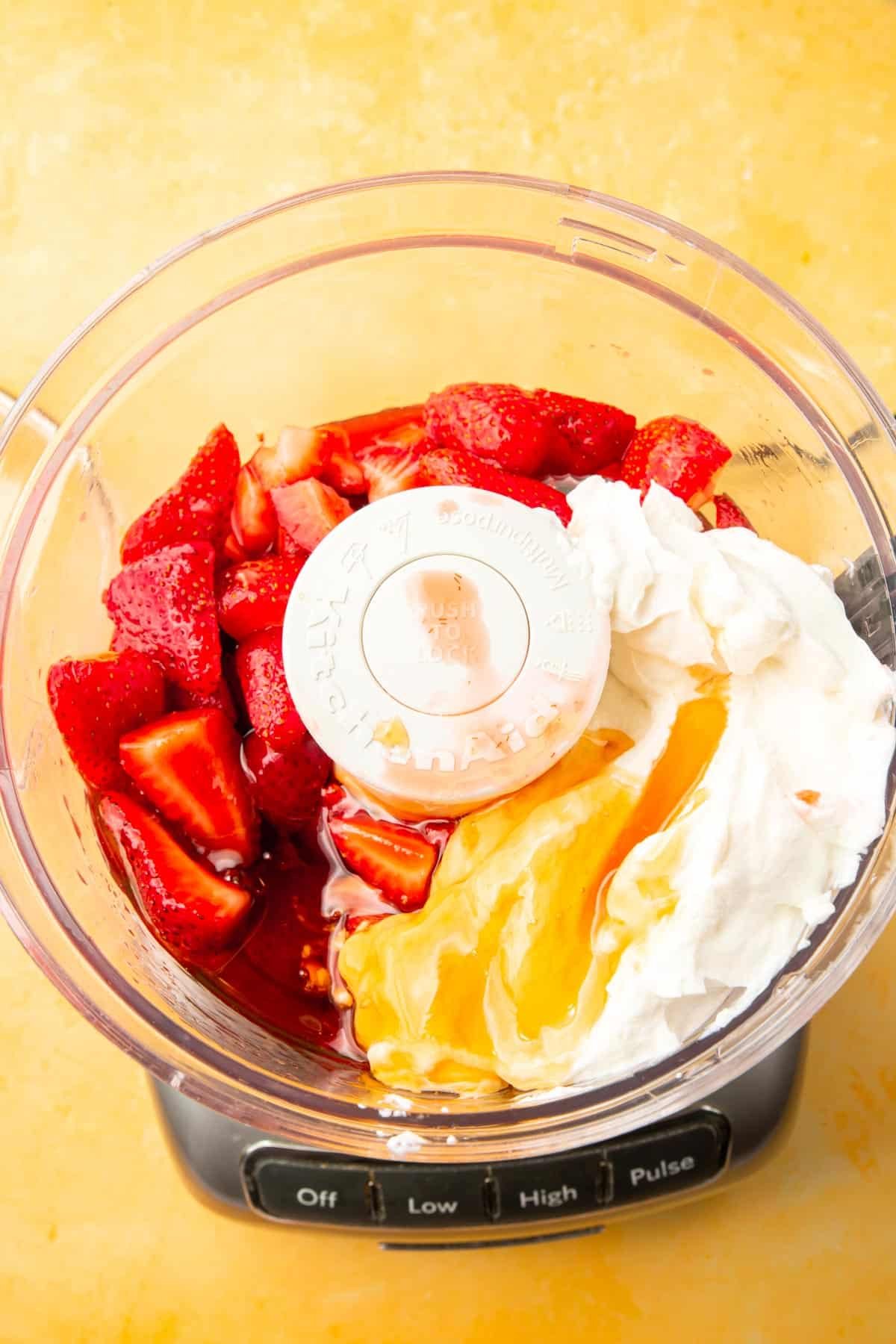 A food processor on a yellow table with greek yogurt, honey, and strawberries in it.