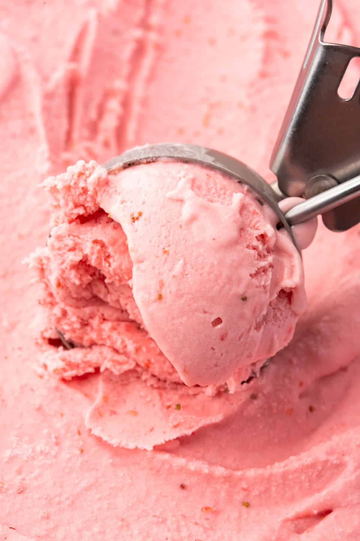 An ice cream scooper scooping strawberry ice cream from a tub.