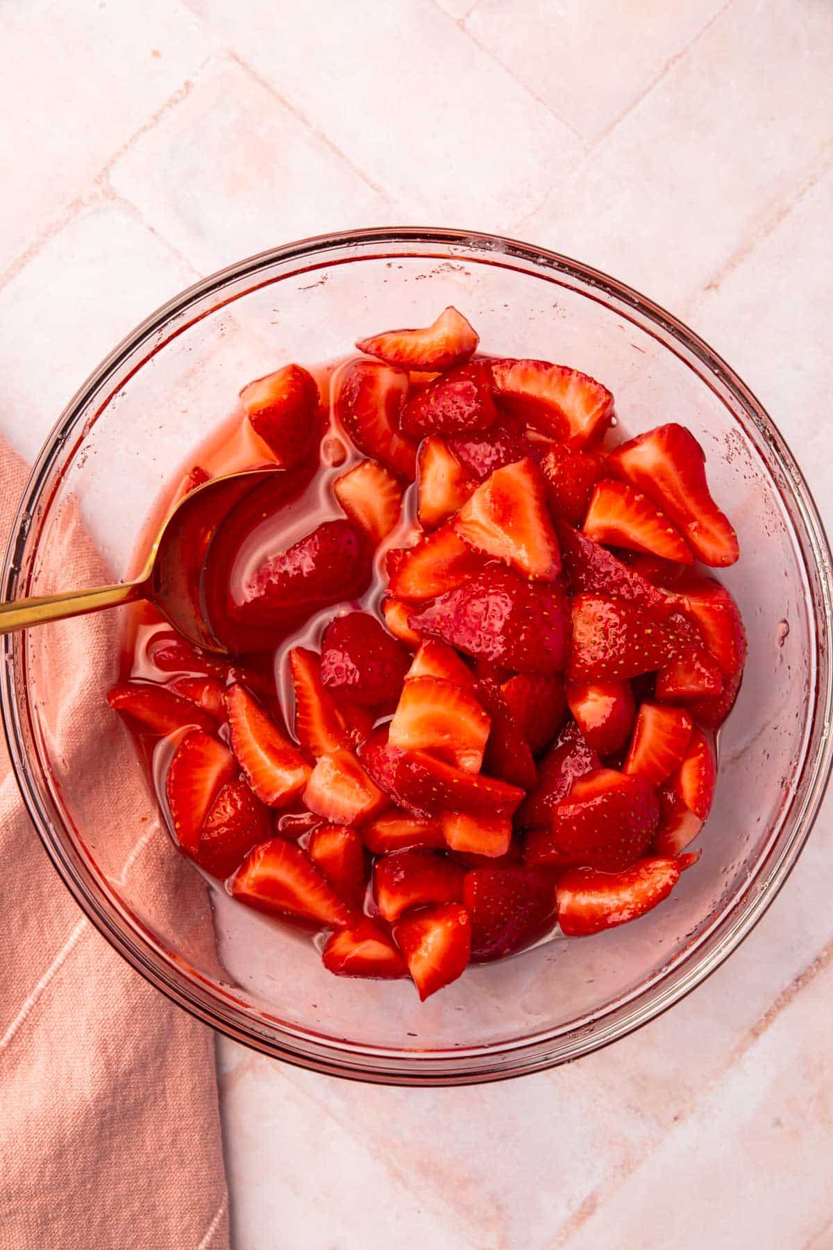 An overhead view of a glass bowl of macerated strawberries with a gold spoon.