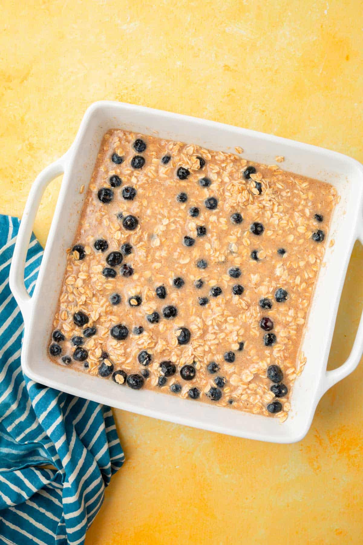 A wet mixture of almond milk, spices, oats, and blueberries in a square baking dish on a yellow table.
