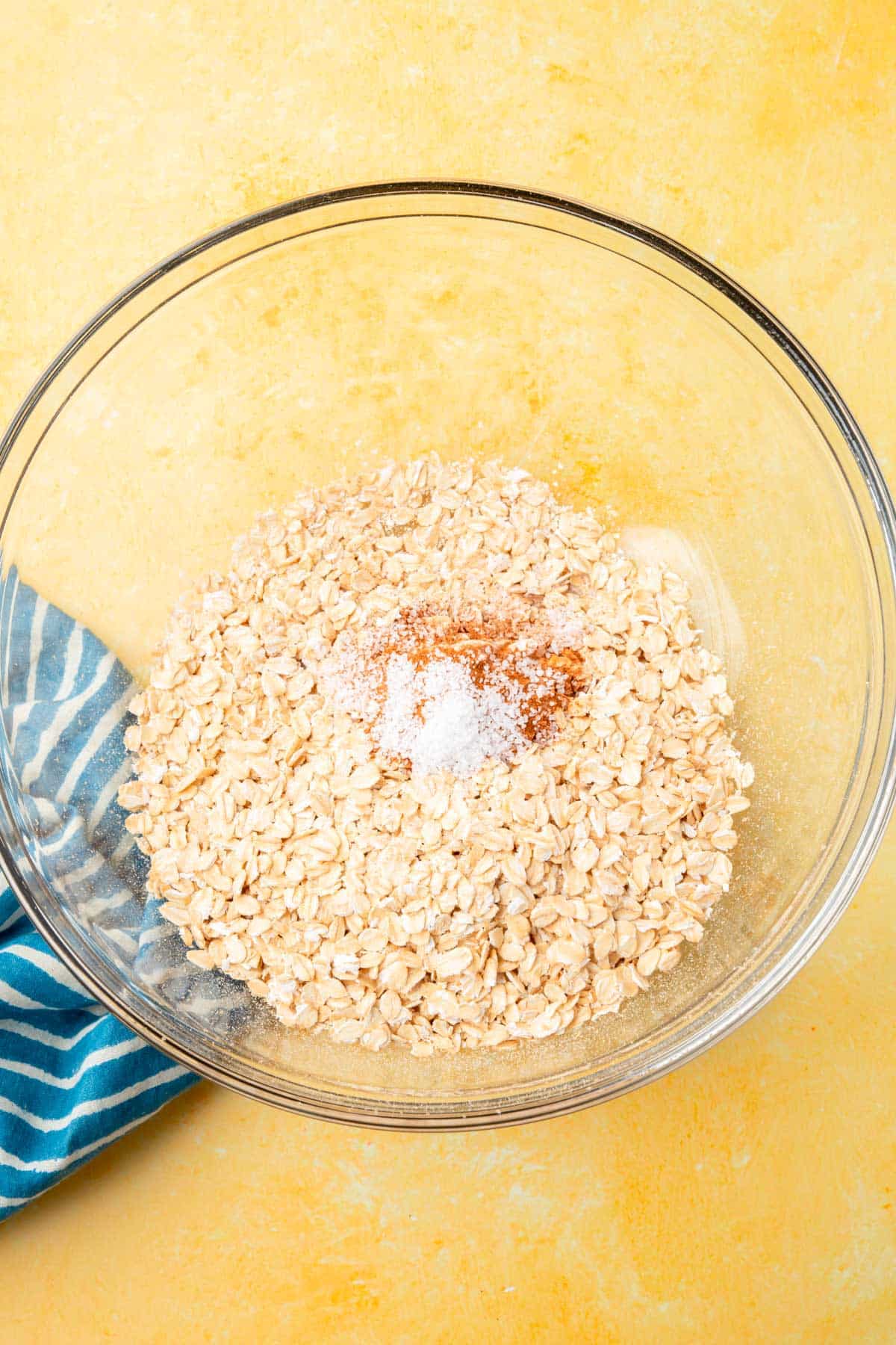 A glass mixing bowl filled with gluten-free oats, salt, baking powder, and cinnamon that has not yet been mixed together.