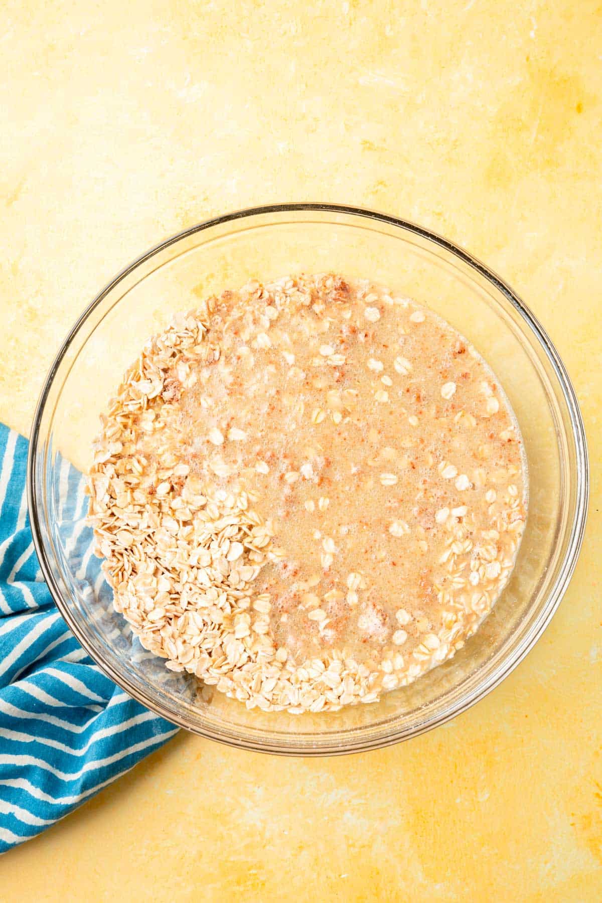 A glass mixing bowl on a yellow table filled with gluten-free oats, cinnamon, and almond milk not yet mixed together.
