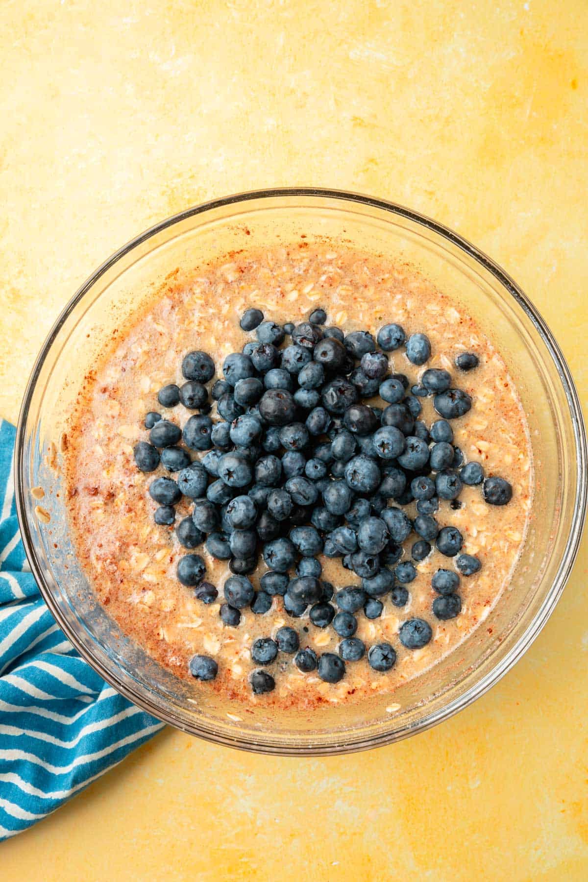 A glass mixing bowl of a wet mixture of oats, almond milk, cinnamon, and applesauce topped with fresh blueberries that have not yet been mixed in.