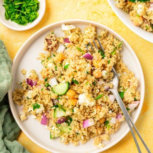 A salad plate with cold quinoa chickpea salad topped with cucumber, onion, and feta with a fork and a bowl of fresh chopped parsley on the side.