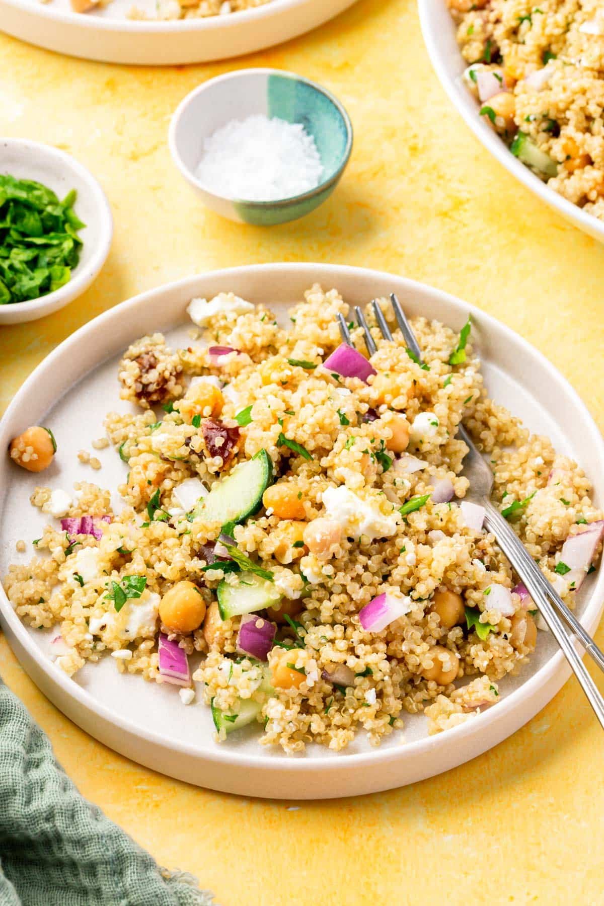 A salad plate with cold quinoa chickpea salad topped with cucumber, onion, and feta with a fork and a bowl of fresh chopped parsley and a bowl of maldon salt on the side.