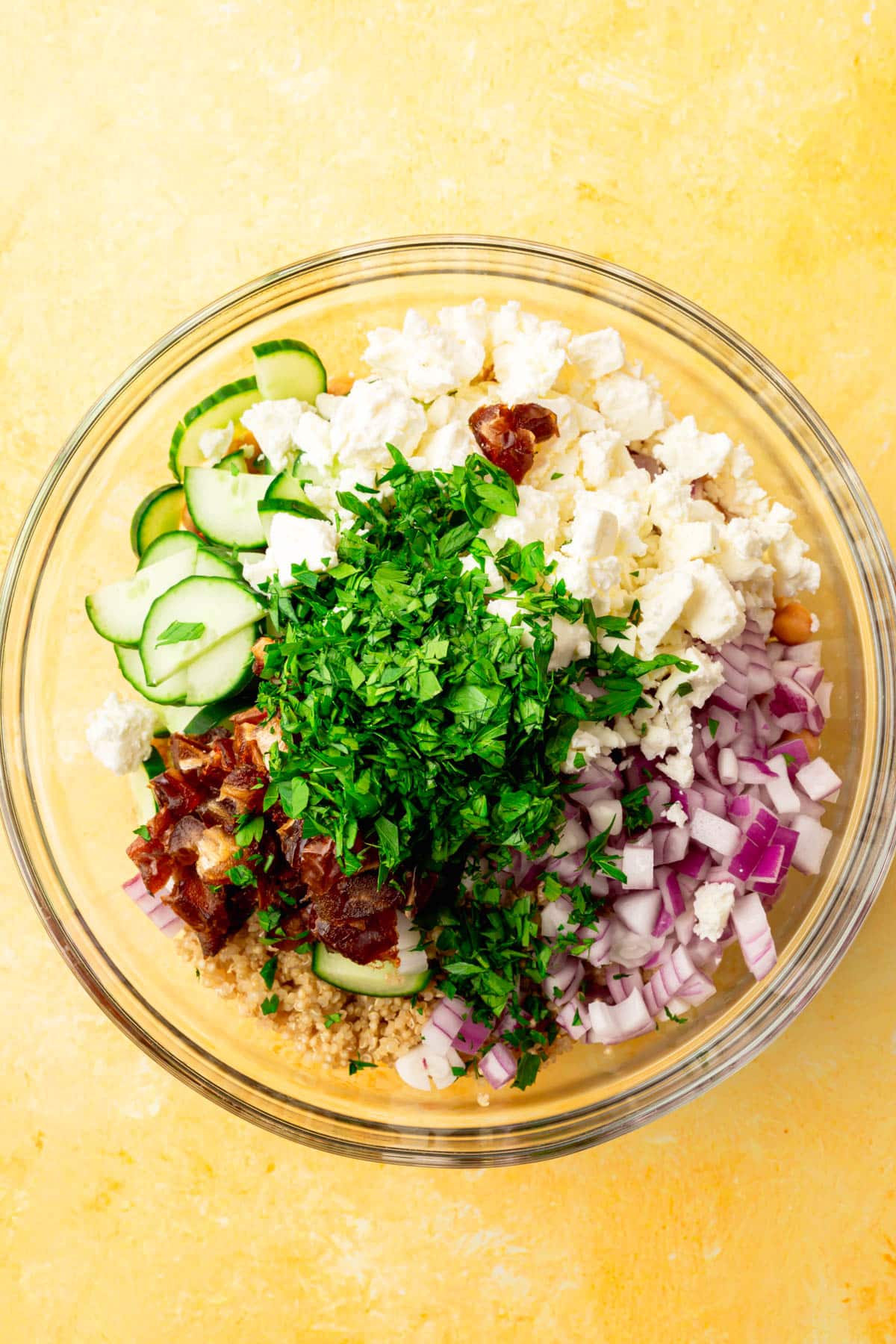 A glass mixing bowl with feta, sliced cucumber, diced red onion, diced dates, and quinoa in it before mixing together.
