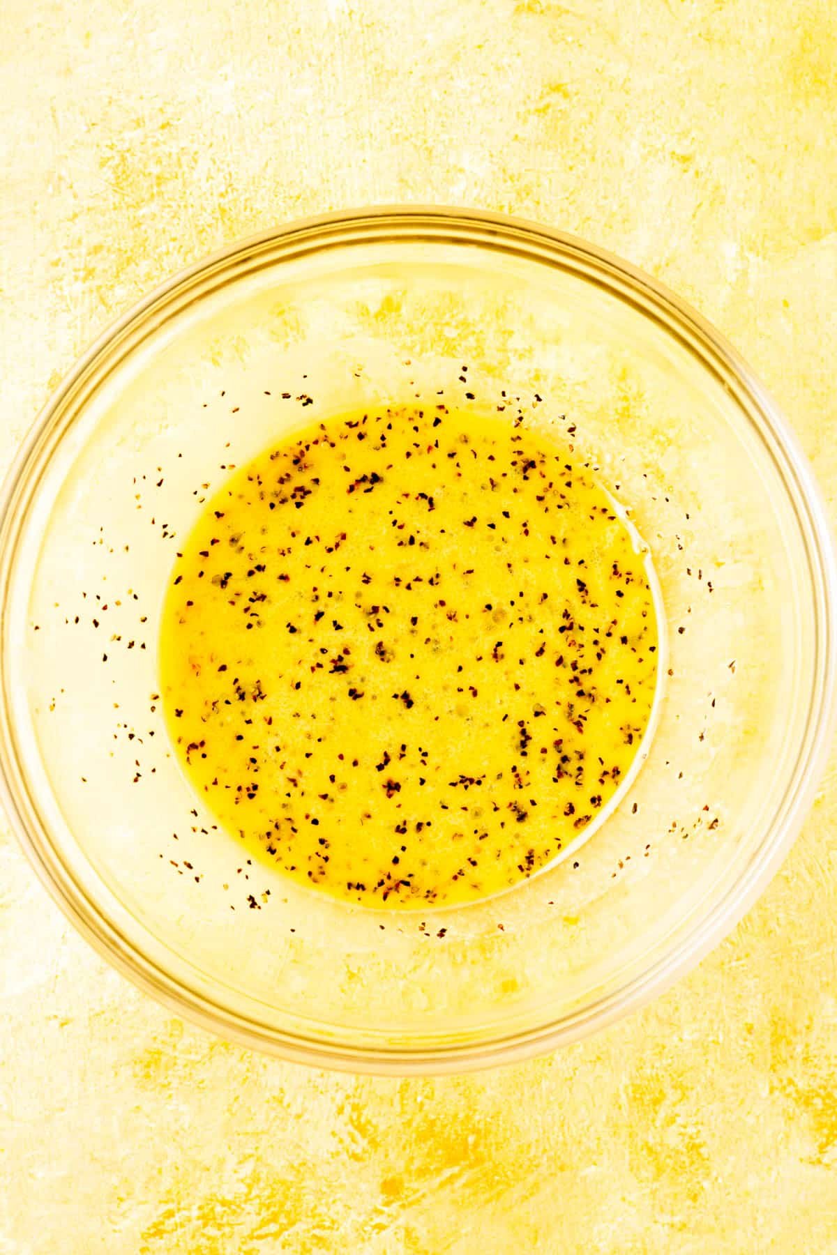 A glass mixing bowl with a lemon juice and olive oil dressing with black pepper in it.