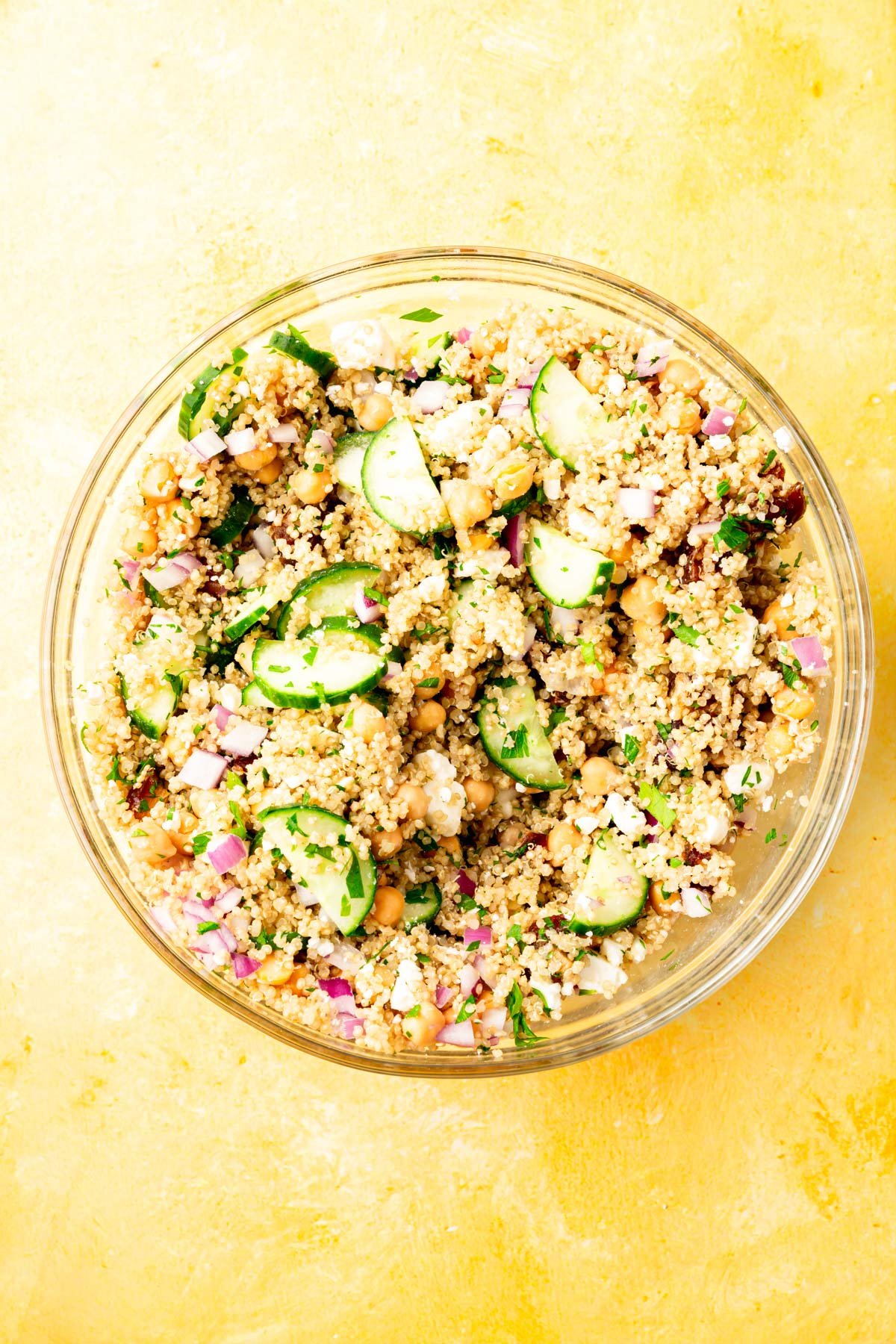 A glass mixing bowl on a yellow table with a chickpea quinoa salad mixed with sliced cucumber, diced red onion, dates, and fresh parsley.